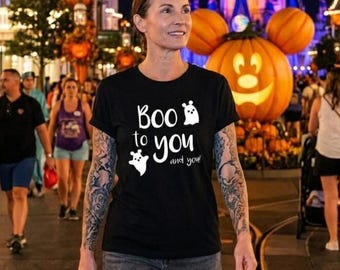Może przedstawiać: Czarny t-shirt z napisem "Boo to you and you!" i białymi grafikami duchów. Osoba nosi koszulkę w świątecznym otoczeniu na zewnątrz, z dekoracjami Halloween i zamkiem w tle.