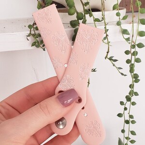 May include: Two pink silicone watch bands with a white floral pattern. The bands have a silver metal button.