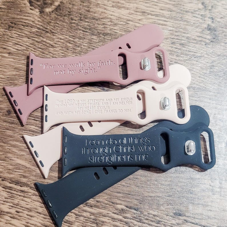 May include: Four different Apple Watch bands with inspirational bible verses printed on them. The bands are in shades of pink and black.