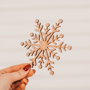 K&ouml;nnte beinhalten: Holz-Schneeflocken-Dekoration mit nat&uuml;rlicher Holzoberfl&auml;che. Die Schneeflocke wird von einer Hand mit rotem Nagellack vor einem wei&szlig;en Hintergrund gehalten. Sie hat sechs symmetrische Spitzen mit aufwendigen Ausschnitten.