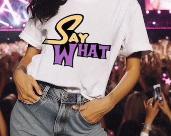 May include: White t-shirt with the words "Say What" in a purple and yellow design. The shirt is paired with blue jeans. The image is taken at a concert with a crowd in the background.