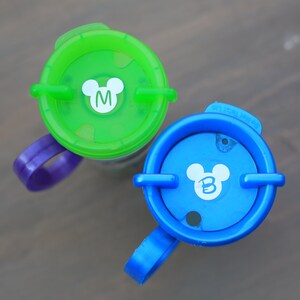 May include: Two colorful plastic cups with Mickey Mouse ears and the letters "M" and "B" on the lids. The cups have purple and blue handles.