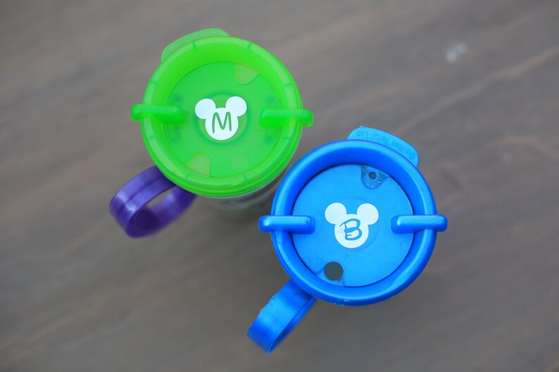 May include: Two colorful plastic cups with Mickey Mouse ears and the letters "M" and "B" on the lids. The cups have purple and blue handles.