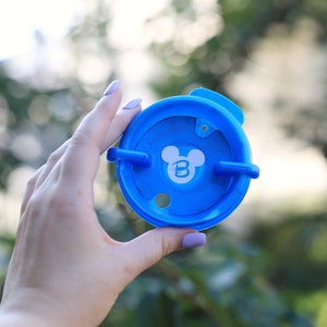 May include: A blue plastic lid with a white Mickey Mouse head and the letter "B" inside the ears. The lid has two tabs on the side.