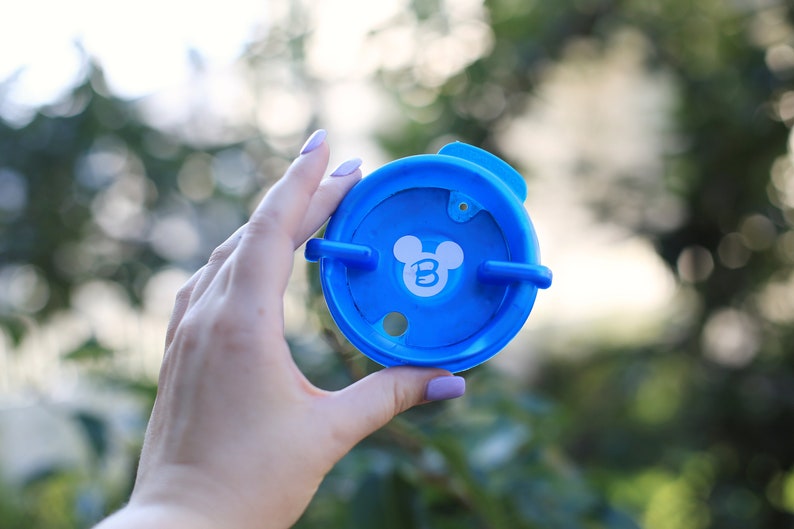 May include: A blue plastic lid with a white Mickey Mouse head and the letter "B" inside the ears. The lid has two tabs on the side.
