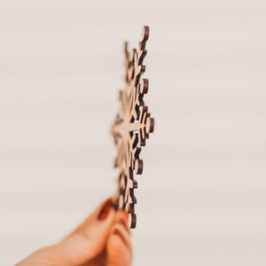 K&ouml;nnte beinhalten: Ein Holz-Schneeflocken-Ornament mit aufwendigen Ausschnitten. Die Schneeflocke ist hellbraun und wird vor einem verschwommenen wei&szlig;en Hintergrund gehalten. Geeignet f&uuml;r Winterdekorationen.