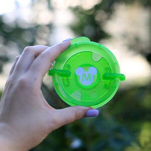 May include: Green plastic lid with a white Mickey Mouse head and the letter "M" in the center. The lid has two tabs on the side.