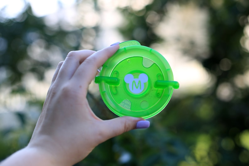 May include: Green plastic lid with a white Mickey Mouse head and the letter "M" in the center. The lid has two tabs on the side.