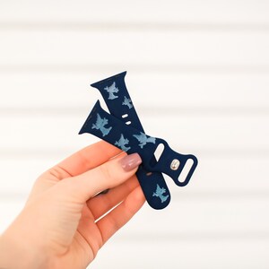 May include: A blue silicone Apple Watch band with a white silhouette pattern of a witch. The band is folded in half and held in a hand.