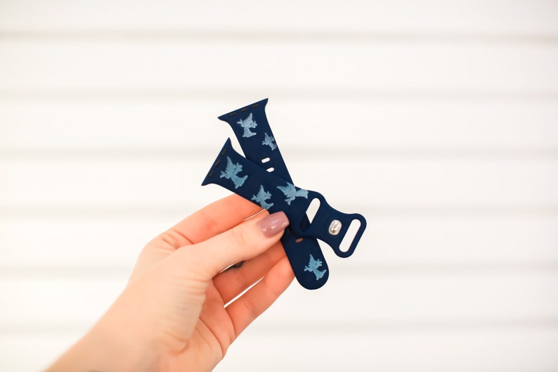 May include: A blue silicone Apple Watch band with a white silhouette pattern of a witch. The band is folded in half and held in a hand.