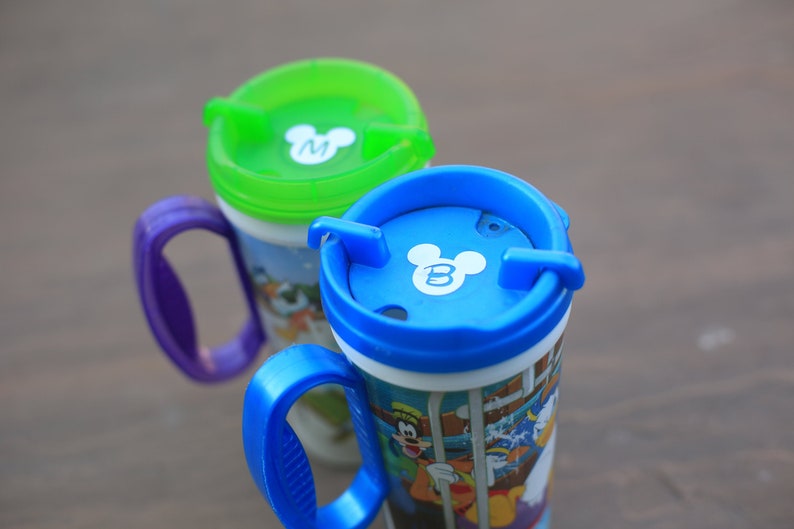 May include: Two colorful Mickey Mouse themed mugs with lids. The mug on the left has a green lid with a white Mickey Mouse head with the letter "M" inside. The mug on the right has a blue lid with a white Mickey Mouse head with the letter "B" inside.