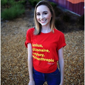 Może przedstawiać: Kobieta w czerwonej koszulce z napisem "Genius, Billionaire, Playboy, Philanthropis - Tony Stark" w ż&oacute;łtych literach. Ma na sobie niebieskie dżinsy i uśmiecha się.