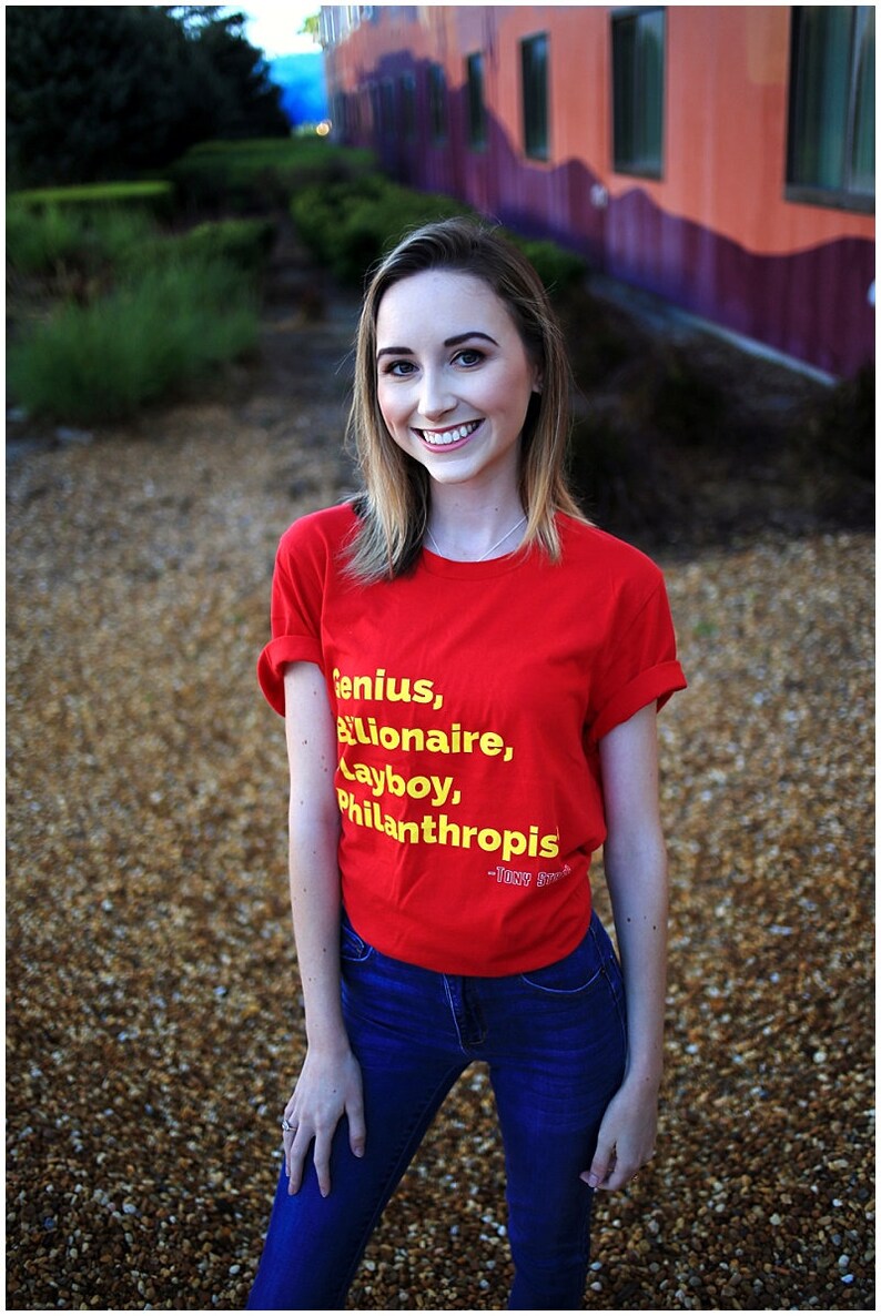 Może przedstawiać: Kobieta w czerwonej koszulce z napisem "Genius, Billionaire, Playboy, Philanthropis - Tony Stark" w ż&oacute;łtych literach. Ma na sobie niebieskie dżinsy i uśmiecha się.