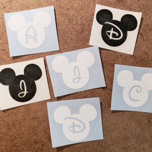 May include: Six Mickey Mouse head stickers with different letters inside. The stickers are on a brown background. The letters are A, J, C, D, and the Disney logo.
