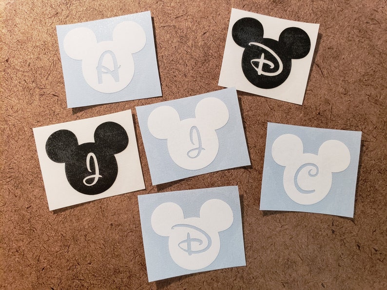 May include: Six Mickey Mouse head stickers with different letters inside. The stickers are on a brown background. The letters are A, J, C, D, and the Disney logo.