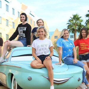 Może przedstawiać: Sześć kobiet pozuje przed kolorowym budynkiem z niebieskim samochodem. Samoch&oacute;d ma kresk&oacute;wkową twarz i jest repliką postaci Złomek McQueen z filmu Cars Disneya Pixara. Kobiety noszą r&oacute;żne koszulki z nadrukiem tekstu. Tekst na koszulkach zawiera "Straight outta dresses", "You and me? We're in a club now", i "Genius, Billionaire, Playboy, Philanthropist".