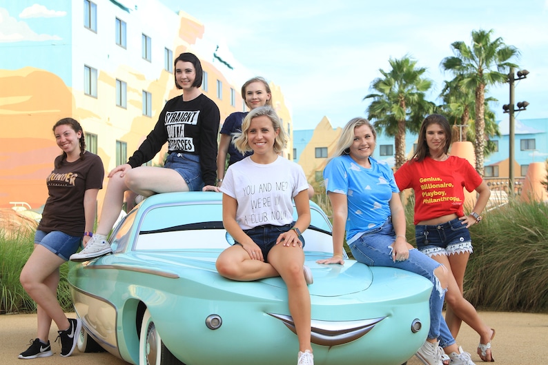 Może przedstawiać: Sześć kobiet pozuje przed kolorowym budynkiem z niebieskim samochodem. Samoch&oacute;d ma kresk&oacute;wkową twarz i jest repliką postaci Złomek McQueen z filmu Cars Disneya Pixara. Kobiety noszą r&oacute;żne koszulki z nadrukiem tekstu. Tekst na koszulkach zawiera "Straight outta dresses", "You and me? We're in a club now", i "Genius, Billionaire, Playboy, Philanthropist".