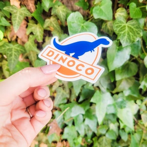 May include: A blue dinosaur sticker with the word "DINOCO" in orange letters on a white background. The sticker is held in a hand against a green leafy background.