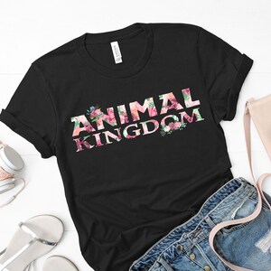 Puede incluir: Camiseta negra con la frase "ANIMAL KINGDOM" en un dise&ntilde;o floral. Las letras est&aacute;n llenas de flores rosas, rojas y verdes. Las mangas est&aacute;n dobladas. Unos auriculares, un bolso y unos vaqueros est&aacute;n junto a la camiseta.