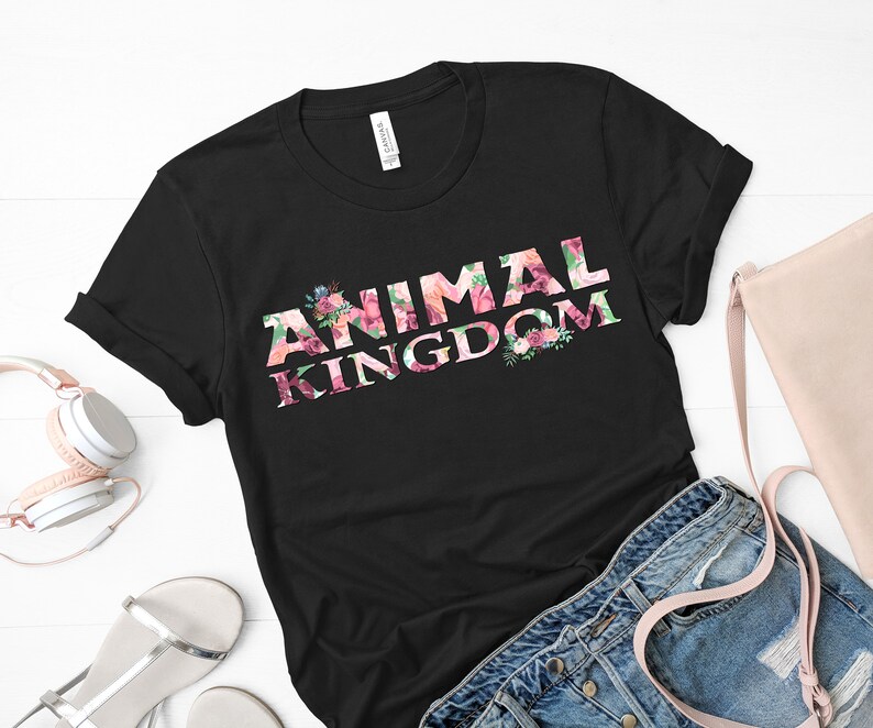 Puede incluir: Camiseta negra con la frase "ANIMAL KINGDOM" en un dise&ntilde;o floral. Las letras est&aacute;n llenas de flores rosas, rojas y verdes. Las mangas est&aacute;n dobladas. Unos auriculares, un bolso y unos vaqueros est&aacute;n junto a la camiseta.