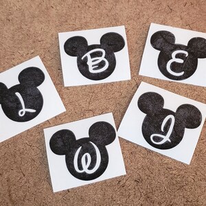 May include: Five black and white Mickey Mouse head stickers with different letters inside each head. The letters are B, E, L, J, and W.
