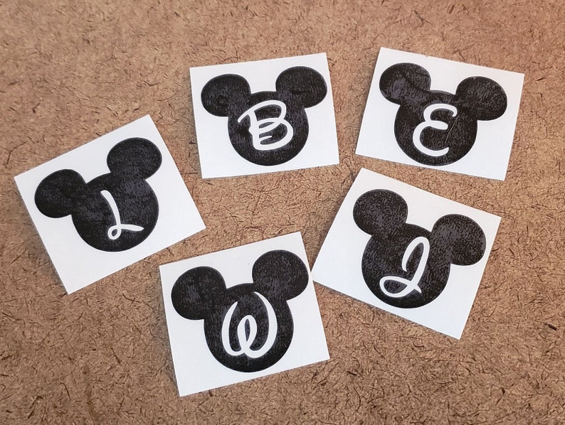 May include: Five black and white Mickey Mouse head stickers with different letters inside each head. The letters are B, E, L, J, and W.