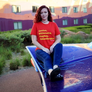 Może przedstawiać: Kobieta w czerwonej koszulce z napisem "Genius, Billionaire, Playboy, Philanthropist" w ż&oacute;łtych literach. Siedzi na fioletowym samochodzie z niebieskimi paskami.