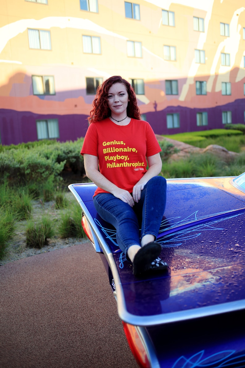 Może przedstawiać: Kobieta w czerwonej koszulce z napisem "Genius, Billionaire, Playboy, Philanthropist" w ż&oacute;łtych literach. Siedzi na fioletowym samochodzie z niebieskimi paskami.