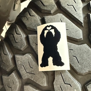 May include: A black Bigfoot silhouette sticker with a heart-shaped hand gesture is placed on a large, textured black tire. The tire has deep grooves and is part of a vehicle, with some of the vehicle's undercarriage visible.