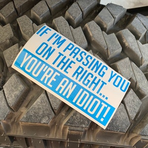 May include: A rectangular sticker with the text "IF I'M PASSING YOU ON THE RIGHT... YOU'RE AN IDIOT!" in white letters on a blue background. The sticker is placed on a black tire with a rugged tread pattern.