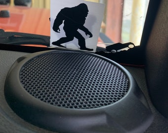 May include: A black speaker with a round, black grill and a black, textured surround. A sticker of a black silhouette of Bigfoot is on the window behind the speaker.