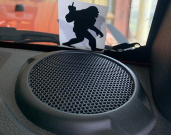May include: A black speaker with a round, black grill and a black, textured surround. A sticker of a black silhouette of a Bigfoot giving the middle finger is on the window behind the speaker.