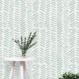 Puede incluir: Una mesa auxiliar blanca con un papel tapiz estampado de color verde claro detrás. Una pequeña planta en maceta se encuentra sobre la mesa y una planta en maceta más grande se encuentra en el suelo en una cesta de alambre.