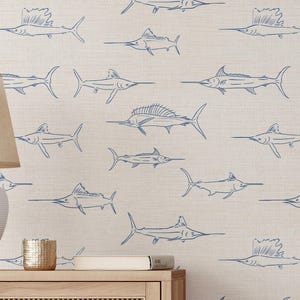 May include: Beige wallpaper with a repeating pattern of blue marlin fish. The fish are drawn in a simple, line-art style. The wallpaper is on a wall behind a white lamp, a wooden cabinet, and other decorative items.