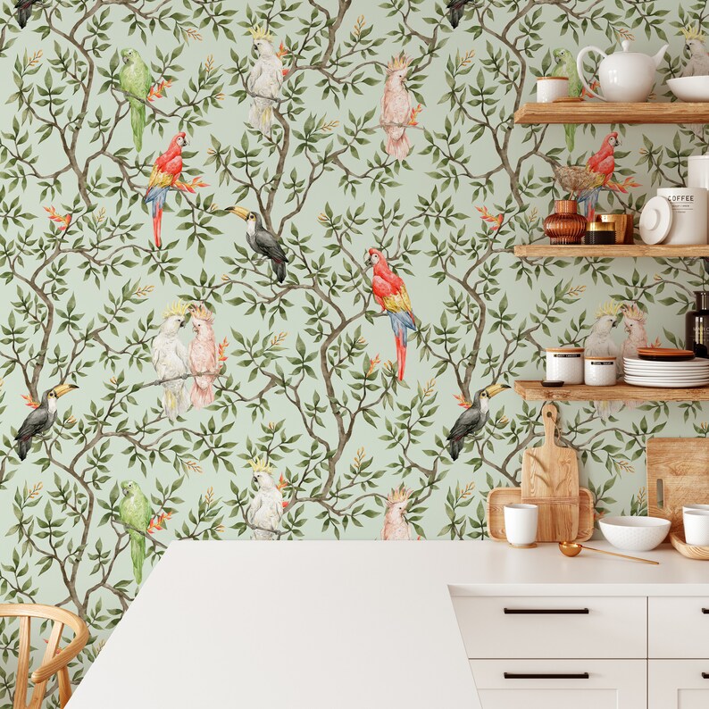 May include: A light green wallpaper featuring a repeating pattern of parrots, toucans, and cockatoos perched on leafy branches. The birds are in shades of red, yellow, green, and white. The wallpaper is in a kitchen setting with white cabinets and wooden shelves.