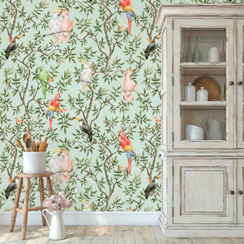 May include: Watercolor wallpaper featuring a repeating pattern of colorful parrots, toucans, and cockatoos perched on leafy branches against a light green background. The design includes a wooden cabinet, a stool, and a white pitcher.
