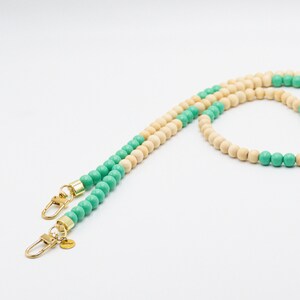 Könnte beinhalten: Ein Perlenriemen mit abwechselnd hellen Holz- und türkisfarbenen Perlen. Der Riemen hat goldfarbene Metallverschlüsse an jedem Ende. Ein kleiner goldener Anhänger hängt an einem Verschluss. Der Riemen befindet sich auf weißem Hintergrund.