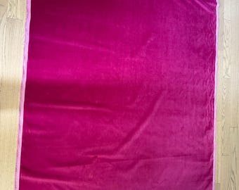 Puede incluir: Un gran trozo rectangular de tela de terciopelo fucsia. La tela tiene una textura suave y afelpada y un color vibrante. Los bordes están acabados con un ribete rosa más claro. La tela está sobre un suelo de madera.