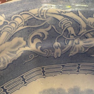May include: A white ceramic bowl featuring a blue transferware pattern. The interior rim displays intricate scrolling floral vines and leaves, while a geometric border separates the upper edge from a central landscape scene with clouds.