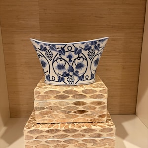 May include: A white ceramic bowl with a blue grape vine and leaf pattern sits atop two stacked rectangular boxes. The boxes feature a textured, light brown, leaf-like inlay pattern across their exterior surfaces, creating a layered display.