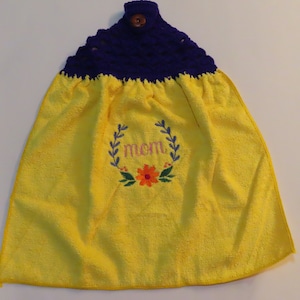 May include: Yellow kitchen towel with a dark blue crocheted top and a wooden button. The towel has the embroidered word "mom" inside a floral wreath design. A decorative and functional kitchen accessory.