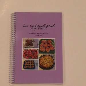 May include: Purple spiral bound cookbook with the title "Low Carb Small Meals Any Time 2" and the subtitle "Certified Health Coach Tina Gill". The cover features photos of various food dishes.
