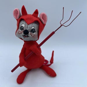 May include: A red felt devil mouse plush toy holding a red metal pitchfork. The mouse has a gray face with black eyes and a black nose. The mouse is wearing a red felt devil costume with a pointed tail.