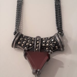Puede incluir: Un collar plateado con un colgante con una piedra triangular de color marrón rojizo. El colgante está enmarcado por un diseño metálico decorativo y texturizado y cuelga de una doble cadena.