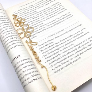 Può includere: Un segnalibro in filo dorato a forma di fiore con la parola "become" scritta in corsivo. Il segnalibro è posizionato su un libro con il testo "Chapter Summary" visibile.