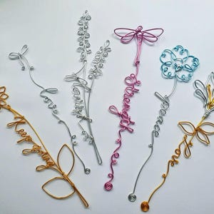 May include: A selection of wire art floral designs in gold, silver, pink, and blue. Each piece features a different flower or insect shape, with words like "Love" and "Peace" incorporated. The pieces are arranged on a white surface. Measurements are in cm.
