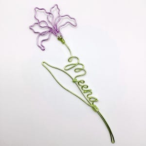 Può includere: Una scultura decorativa in filo metallico a forma di fiore con petali viola e uno stelo verde. Lo stelo è formato in scrittura corsiva con il nome Meena, integrato nella struttura della foglia e del gambo, creando un pezzo unico di filo.