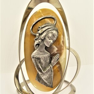 May include: A silver metal frame with a yellow and white oval-shaped stone featuring a silver metal image of a woman with her hands clasped in prayer.