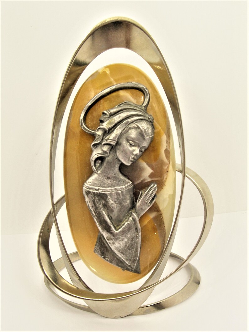 May include: A silver metal frame with a yellow and white oval-shaped stone featuring a silver metal image of a woman with her hands clasped in prayer.
