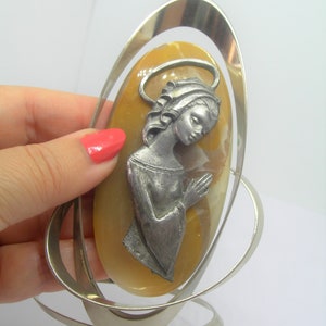May include: A silver oval frame with a yellow oval stone inside. The stone has a silver figure of a woman with her hands clasped in prayer.